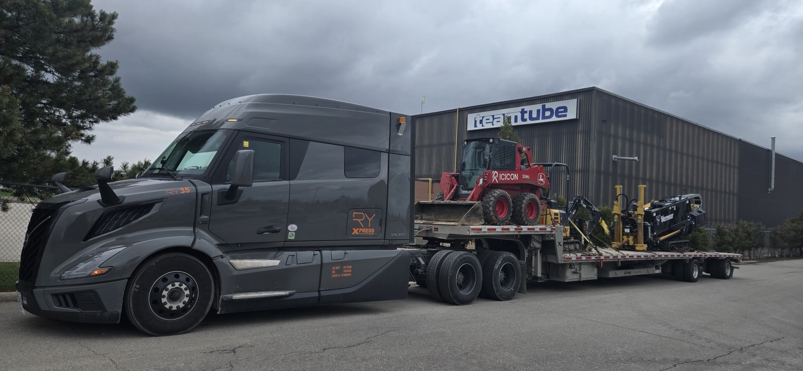 Gray RY Volvo hauling skid steer and equipment on flatbed