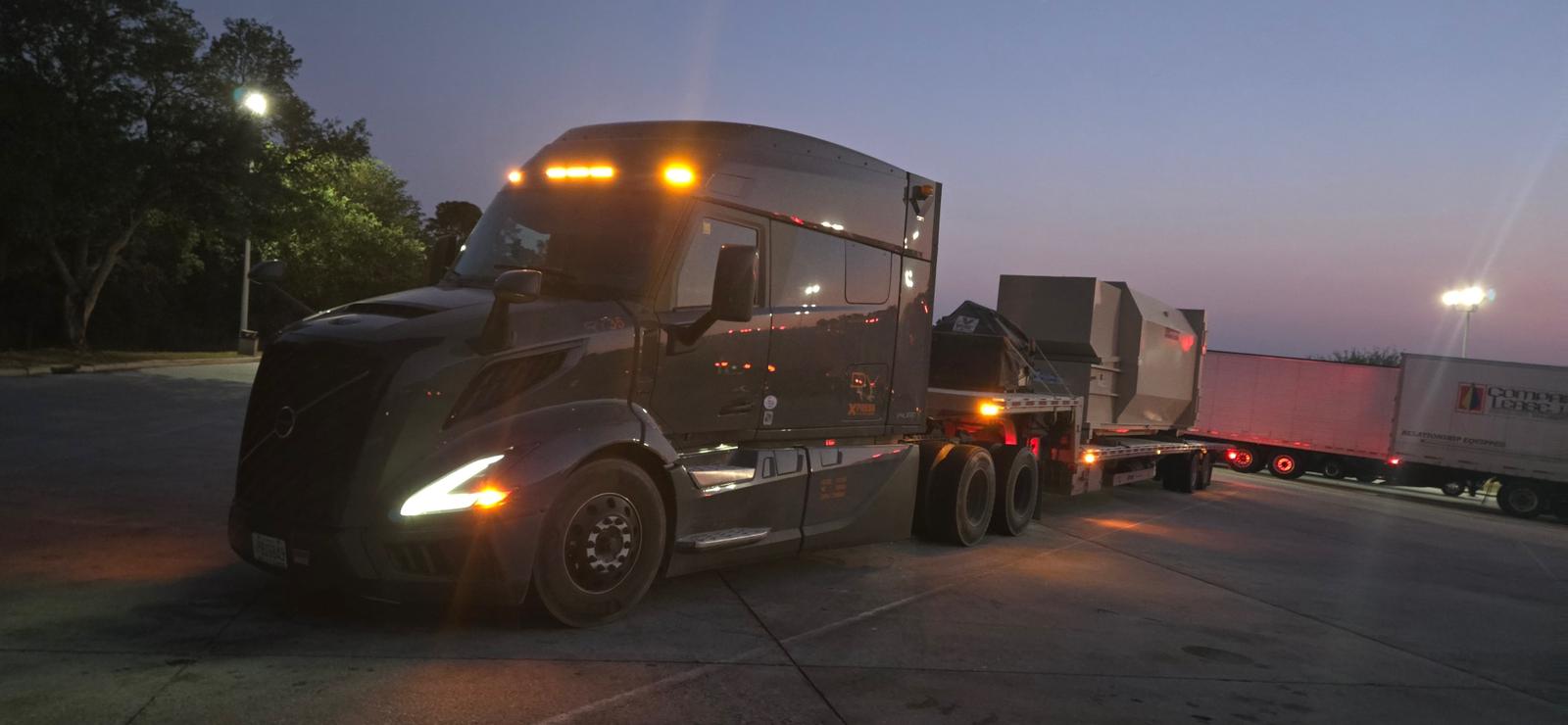 Gray Volvo VNL with amber lights hauling industrial equipment at dusk