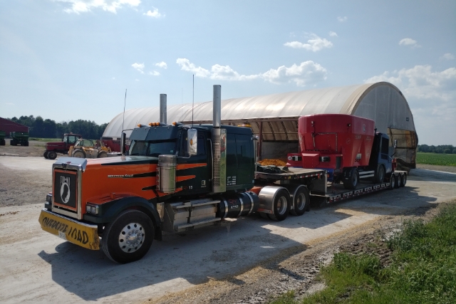 Orange and black Western Star hauling red truck on lowboy in rural setting